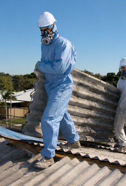 Two operatives in full PPE carefully carrying a corrugated asbestos cement roof sheet across a residential roof in kent
