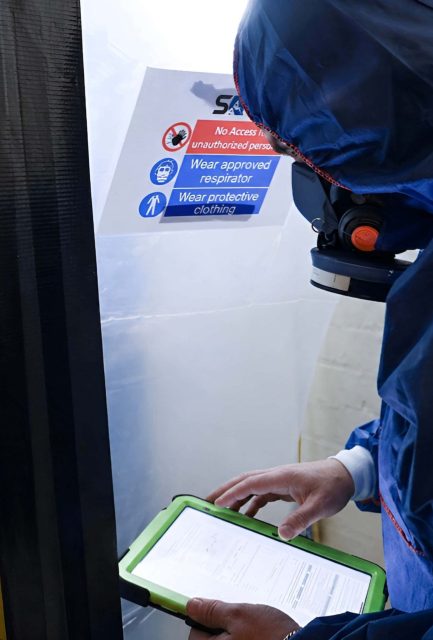 Surrey Asbestos operative in full PPE reviewing documentation at a sealed asbestos hazard enclosure entry point in Surrey
