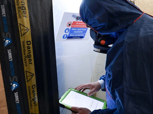Surrey Asbestos operative in full PPE reviewing documentation at a sealed asbestos hazard enclosure entry point in Surrey