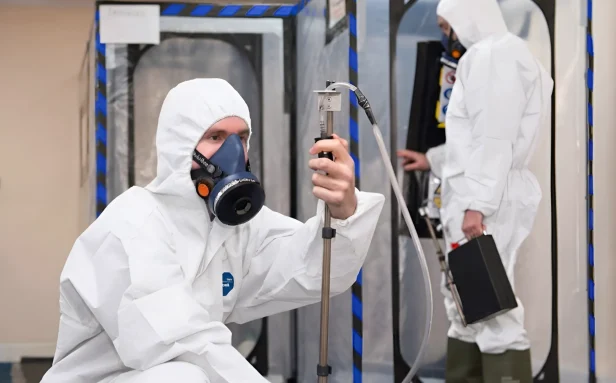 Asbestos analyst in full PPE setting up air monitoring equipment beside a sealed enclosure on a Surrey site.